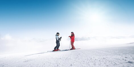 Two skiers stand on freshly groomed slope in Ski amadé overlooking a foggy valley in bright sunlight